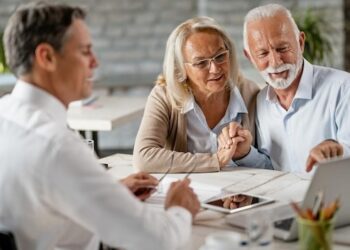 Happy senior couple holding hands and using laptop while having a meeting with financial advisor in the office Senior man is pointing at something on laptop