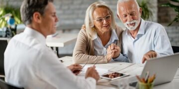 Happy senior couple holding hands and using laptop while having a meeting with financial advisor in the office Senior man is pointing at something on laptop