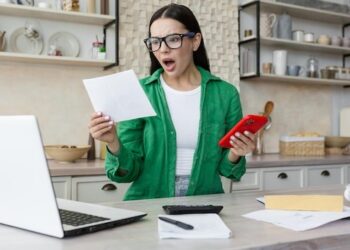 Shocked young woman reading a received letter while sitting in the kitchen at home he holds the