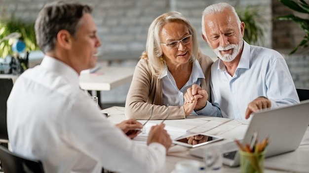 Happy senior couple holding hands and using laptop while having a meeting with financial advisor in the office Senior man is pointing at something on laptop